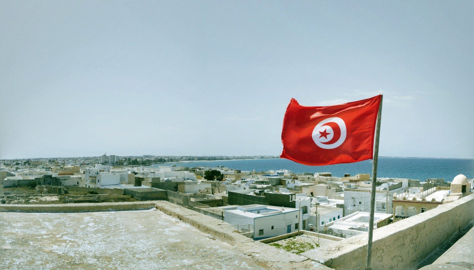 A vibrant Tunisian flag waving over the coastal town of Mahdia with a waterfront view.